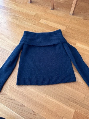 Mörkblå off shoulder stickad tröja - Säljer en mörkblå stickad tröja med bred off shoulder-krage och långa ärmar. Tröjan har ribbstickade muddar vid ärmslut och nederkant, vilket ger en snygg och avslappnad look. Perfekt för dig som gillar trendiga och bekväma plagg.
