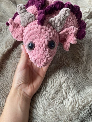 Rosa virkad dinosaurie mjukisdjur - Supergullig virkad dinosaurie i rosa med lila taggar och grå horn. Tillverkad i mjukt garn och har stora svarta ögon. Perfekt som dekoration eller mysig kompis att krama. Unik och handgjord!