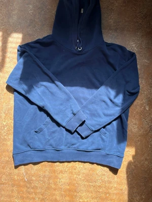 Mörkblå hoodie med huva - En enkel och stilren mörkblå hoodie med huva och dragsko. Hoodien har en klassisk känguruficka framtill och ribbade muddar vid ärmslut och nederkant. Perfekt för en avslappnad och bekväm look.