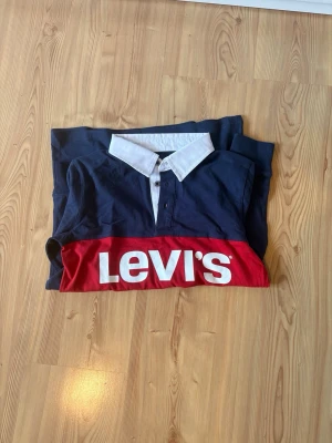 Blå långärmad Levi's rugby tröja - Säljer en snygg långärmad rugby tröja från Levi's i marinblått med vit krage och en bred röd panel över bröstet med stor vit Levi's-logga. Tröjan har knappar vid halsen och är gjord i mjuk bomull. Perfekt för dig som gillar streetwear och klassisk stil.