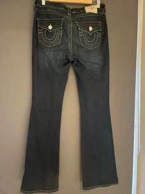 Mörkblå bootcut jeans med kontrastsömmar - Säljer ett par mörkblå bootcut jeans från true religion med snygga fickdetaljer bak. Helt oanvända och i storlek 29 så det passar båda M och L 