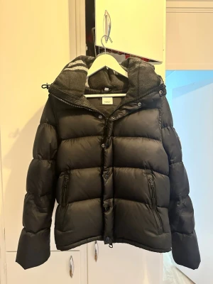 Svart dunjacka med huva - Snygg och svart Burberry dunjacka med stor vadderad huva och dragkedja framtill. Jackan har två dragkedjefickor, quiltad design och ribbade muddar. Perfekt för kalla dagar och har  med detaljer på huvan. Jag säljer den eftersom att jag inte använder den. Har ni några frågor är det bara att fråga, pris kan diskuteras😁