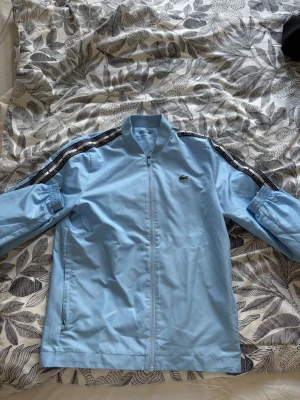 Ljusblått tracksuit set Lacoste - Säljer ett ljusblått tracksuit set från Lacoste med dragkedja på jackan och resår i midjan på byxorna. Setet har svarta och vita detaljer längs ärmarna och byxbenen samt den klassiska Lacoste-loggan på bröstet och höften. Perfekt för en sportig och avslappnad stil.