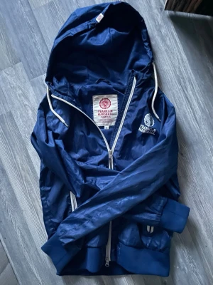 Blå vindjacka från Franklin & Marshall - Snygg blå vindjacka från Franklin & Marshall med vit dragkedja och vita snören i huvan💙🤍💫.                          OBS! Skulle säga att den sitter som en S och inte M i storleken.                    Jackan har ribbade muddar, två fickor med dragkedja och logga på bröstet. Tillverkad i lätt, glansigt material som står emot vind. Vill tillägga att vänstra dragkedjan är ur funktion