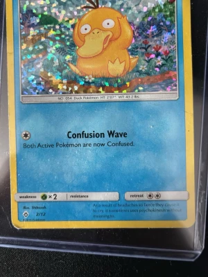Holo Psyduck Pokémonkort - Donken 2018 Säljer ett holografiskt Psyduck Pokémonkort med färgglad och glittrig framsida. Kortet har blå och gul ram, motiv av Psyduck och attacken Confusion Wave. Baksidan har klassiskt Pokémon-mönster med Pokéball. Perfekt för samlare eller Pokémonfans.