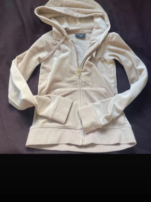 Beige velour hoodie med dragkedja - Snygg beige hoodie i mjuk velour med dragkedja framtill och kängurufickor. Har huva med snören och ribbade muddar vid ärmar och nederkant. Perfekt för en chill och avslappnad stil.