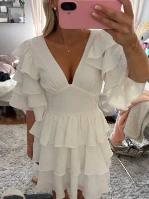 Vit volangklänning med v-ringning - Supersöt vit klänning med flera lager volanger och v-ringad hals. Klänningen har korta, voluminösa ärmar och en figurnära midja som ger en feminin siluett. Perfekt för sommarens alla tillfällen!     Nypris: 899kr