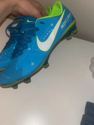 Nike Mercurial blå fotbollsskor - Säljer ett par Nike Mercurial fotbollsskor i blått med vita och neongröna detaljer. Skorna har snörning, låg profil och dobbar för spel på gräs. Swoosh-logga på sidan och Mercurial-tryck. Insidan är neongrön och yttermaterialet är syntetiskt.
