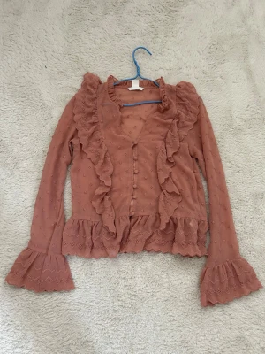 Rosa volangblus från H&M - Supersöt rosa blus från H&M med volanger längs knäppningen och ärmsluten. Blusen har diskret broderat mönster, genomskinligt tyg och långa ärmar med volangdetaljer. Perfekt för dig som gillar romantisk och trendig stil.