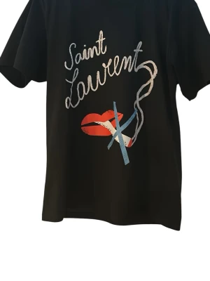 Svart Saint Laurent t-shirt med tryck - Svart t-shirt från Saint Laurent med coolt tryck framtill som visar röda läppar, cigarett och texten 'Saint Laurent'. Klassisk passform och korta ärmar. Tillverkad i mjuk bomull för skön känsla och stilren look.