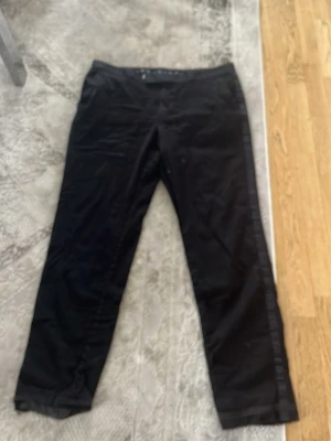 Svarta raka byxor med sidodetalj - Säljer ett par svarta byxor med rak passform och diskret sidodetalj längs benen. Byxorna har enkel midja och är tillverkade i ett mjukt tyg som ger en clean look. Perfekta för dig som gillar minimalistisk stil.