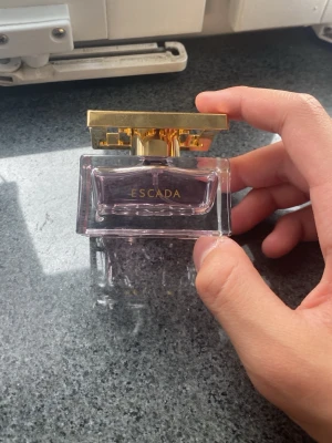 Escada parfym med guldigt lock - Snygg parfym från Escada i en genomskinlig glasflaska med lila toner och ett lyxigt, guldigt lock. Flaskan har en elegant och modern design som känns exklusiv och passar perfekt på sminkbordet.Aldrig använt. Ny pris 699. Skriv till mig för frågor och bud. 
