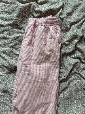 Randiga rosa pyjamasbyxor - Mysiga pyjamasbyxor i ljusrosa och vitt med tunna vertikala ränder. Byxorna har resår i midjan med dragsko och sidofickor. Tillverkade i ett mjukt och lätt tyg som passar perfekt för avslappnade kvällar hemma.