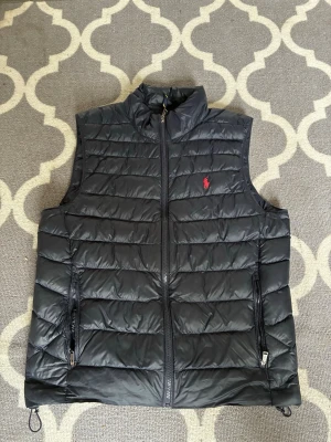 Svart dunväst från Polo Ralph Lauren - Snygg svart dunväst från Polo Ralph Lauren med quiltad design och dragkedja framtill. Västen har två fickor med dragkedja och den klassiska röda loggan broderad på bröstet. Perfekt för lager på lager och stilren look.