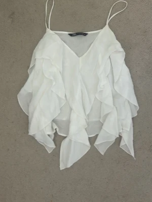 Vit volangig blus - Säljer en superfin vit linne blus med tunna axelband och v-ringning. Toppen har flera lager av volanger som ger en flowy och luftig känsla. Perfekt för varma dagar och ger en riktigt söt och trendig look.