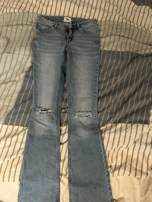 Bootcut jeans med slitningar från 157 - Oanvända helt nya snygga ljusblå bootcut jeans från 157 med slitningar över knäna. Klassisk femficksmodell och låg midja. Perfekta för dig som gillar en avslappnad och trendig stil. Tillverkade i denim. Affärs pris 400kr