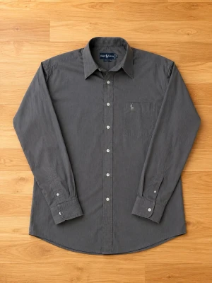 Grå skjorta från Ralph Lauren - Stilren mörkgrå skjorta från Ralph Lauren med klassisk krage och vita knappar. Skjortan har en bröstficka med broderad logga och är tillverkad i mjuk bomull. Perfekt för dig som gillar en clean och tidlös look.