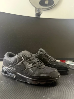 Svarta Nike Air Max 90 sneakers - Säljer ett par helsvarta Nike Air Max 90 sneakers med klassisk siluett. Skorna har ovandel i mesh och läder, synlig Air Max-dämpning i sulan och diskreta Nike-loggor på sidan och plösen. Perfekta för dig som gillar en clean och sportig look.