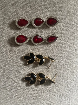 Två par statementörhängen i rött och svart - Säljer två par snygga örhängen. Ett par med droppformade röda stenar omgivna av små klara stenar i silverfärg, och ett par med svarta bladformade stenar i guldfärgad infattning. Perfekta för dig som vill sticka ut med accessoarerna!