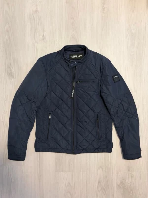 Replay Jacka - Snygg mörkblå quiltad jacka från Replay med dragkedja framtill, tre framfickor med dragkedja och ståkrage med tryckknapp. Jackan har långärmad design och diskreta detaljer på ärmar och axlar. Perfekt till höst och vår. Storlek: S