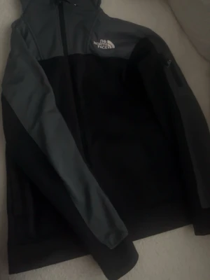 Svart och grå vindjacka The North Face - Snygg vindjacka från The North Face i svart och mörkgrått. Jackan har dragkedja framtill, hög krage och logga på bröstet. Tillverkad i slitstarkt material och har en ficka på ärmen. Perfekt för blåsiga dagar.