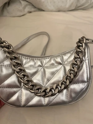 Silvrig quiltad handväska med kedja - Trendig handväska i metallic-silver med quiltat mönster och en chunky silvrig kedja som axelrem. Väskan har en halvmåneform och är tillverkad i glansigt konstläder. Perfekt accessoar för att lyfta din outfit.