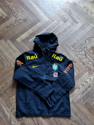 Svart Brasilien Windbreaker - Svart Brasilien Windbreaker i storlek M. Har aldrig använts och är i perfekt skick. Kvaliteten är suverän och den är perfekt för fotbolls älskare som vill vara redo inför dem kommande årstiderna