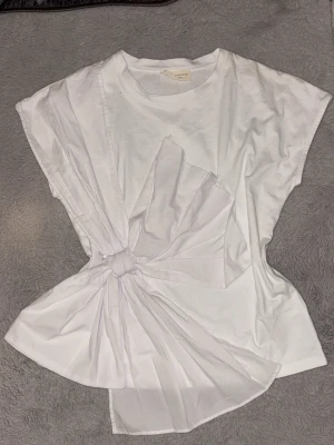 Vit topp med rosett  - Säljer en unik vit topp med korta ärmar och en stor, iögonfallande knutdetalj framtill. Toppen har asymmetrisk design och är gjord i mjukt bomullstyg med extra tygparti som ger volym och cool effekt. Perfekt för dig som vill sticka ut med din stil. Köpt i Grekland aldrig använd