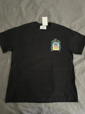 Svart Casablanca t-shirt med tryck - Svart t-shirt från Casablanca med färgglatt tryck av ett fönster och texten 'Avenida Casablanca' både fram och bak. Klassisk passform och rund halsringning. Perfekt för dig som vill ha en stilren men ändå unik look.
