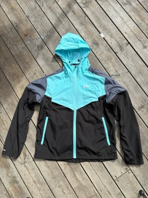 Nike turkos och svart vindjacka - Snygg vindjacka från Nike i turkos, svart och grått. Jackan har huva, hel dragkedja framtill och två fickor med turkosa dragkedjor. Lätt material som passar perfekt för blåsiga dagar. Nike-logga på bröstet och elastiska muddar vid ärmsluten.Passar storlek xs-s