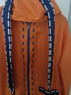 Naruto Uzumaki cosplayjacka orange - Cosplayjacka inspirerad av Naruto Uzumaki från anime-serien Naruto. Jackan är orange med blå och vita detaljer, dragkedja framtill och huva. Långa ärmar och ribbade muddar. Perfekt för cosplay eller maskerad.