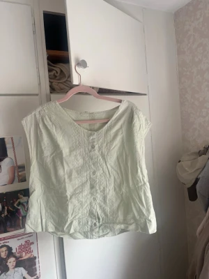 Ljusgrön blus med knappar - Söt ljusgrön blus med v-ringning och dekorativa knappar framtill. Blusen har korta ärmar och fina broderade detaljer som ger en romantisk känsla. Perfekt till sommaren och enkel att matcha med jeans eller kjol.