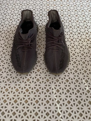 Mörkgrå yeezys  - Säljer ett par mörkgrå sneakers med stickad ovandel och rund tå. Skorna har elastiska snören och en ribbad sula i beige och grå toner. Perfekta för dig som gillar en sportig och modern look. Passar till det mesta och är riktigt bekväma.