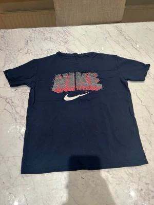 Mörkblå Nike t-shirt med tryck - Snygg mörkblå t-shirt från Nike med ett stort grafiskt tryck i rött, vitt och grått på bröstet. Klassisk passform och rund halsringning. Perfekt för dig som gillar sportig stil och vill ha något bekvämt till vardags.