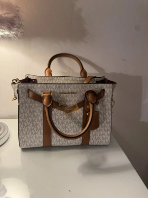 Michael Kors beige handväska - Snygg beige handväska från Michael Kors med MK-logga över hela väskan. Väskan har bruna detaljer, guldfärgade metalldelar och både handtag och axelrem. Invändigt finns flera fack och dragkedja. Perfekt för dig som gillar stilrena accessoarer.