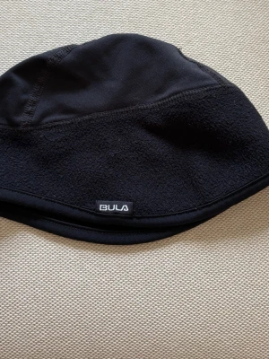 Svart fleecehatt från Bula - Snygg svart hatt från Bula i mjuk fleece och slät textil upptill. Klassisk bucket hat-modell med diskret Bula-logga framtill. Perfekt för kalla dagar när du vill hålla stilen och värmen. Kolla gärna mina andra annonser ❗️
