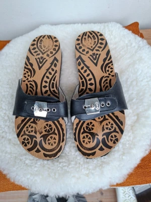 Scholl Pescura Flat Boho läder och trä strlk 37 - Anatomiska sandaler i svart läder som är vadderade invändigt för god komfort med justerbart silvermetallspänne och bottensulor i bl a. bokträ (ingen svettlukt ) 🙂. Passar en fot med 23,3-23,5 cm längd. Varsamt använda, Några slitningar på den svarta yttersulan, men de behöver inte sulas om i det skick de är nu.