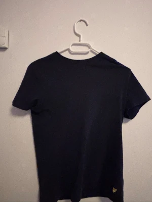 Svart t-shirt från Lyle & Scott - En stilren svart t-shirt från Lyle & Scott Junior med klassisk passform och korta ärmar. T-shirten har en liten gul logga nertill och är gjord i mjuk bomull, perfekt för en clean och enkel look.