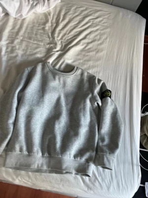 Grå stone island sweatshirt - Snygg grå sweatshirt i mjukt material med rund hals och ribbade muddar. Tröjan har en svart patch med gul och grön logotyp på vänster ärm som ger en cool detalj. Perfekt till jeans eller joggers för en avslappnad look.