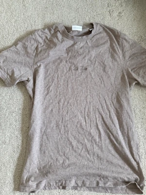 Beige t-shirt från Les Deux - Säljer en stilren beige t-shirt från Les Deux med diskret broderad logga på bröstet. T-shirten har rund halsringning och korta ärmar. Tillverkad i mjuk bomull för en skön känsla och enkel att matcha till olika outfits.