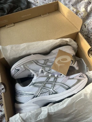 Vita Asics sneakers med silverdetaljer - Säljer ett par fräscha vita Asics sneakers med silvriga detaljer och mesh-överdel. Skorna har en sportig look med klassisk Asics-logga på sidan och vita snören. Perfekta för dig som gillar stilrena och bekväma sneakers.