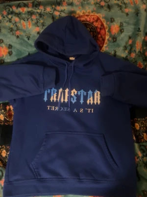 Blå Trapstar hoodie med tryck - Säljer en blå hoodie från Trapstar med vit och blå text på bröstet där det står Trapstar IT'S A SECRET'. Hoodien har huva med dragsko och en stor magficka. Perfekt för dig som gillar streetwear och vill ha en skön och stilren look. Funkar på M 