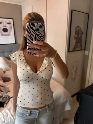 Vit prickig omlottopp med volang - Söt vit omlottopp med svarta prickar och volangdetaljer vid axlarna. Djup v-ringning och ärmlös design ger en fräsch och somrig look. Perfekt att matcha med jeans eller kjol för en trendig outfit.