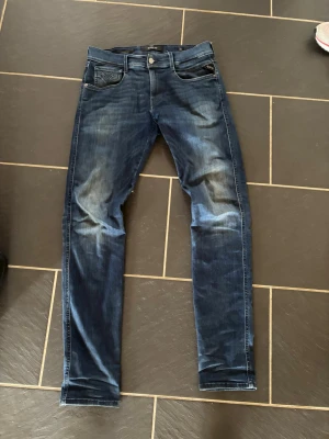 Replay anbass jeans  - Snygga mörkblå slim fit jeans med slitningar och tvättade detaljer. Klassisk femficksmodell med dragkedja och knapp i midjan. Jeansen har raka ben och är tillverkade i stretchigt denimtyg för extra komfort. W30 L34