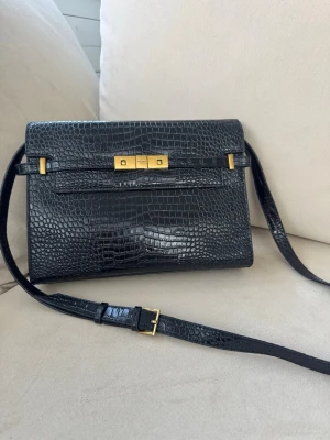 Svart Saint Laurent handväska i läder - Snygg svart handväska från Saint Laurent i croc print med justerbar axelrem och guldfärgade metalldetaljer. Väskan har ett elegant lås framtill och innerfack med dragkedja. Inköpt i Spanien och har aldrig blivit använt.