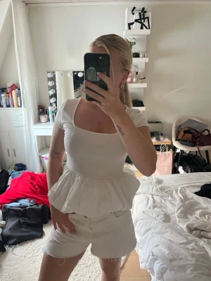 Vit volangblus med kort ärm - Säljer en söt vit blus med volang nedtill och korta ärmar. Toppen har en rundad, något djupare urringning och är figurnära upptill. Perfekt för dig som gillar en feminin och trendig stil.
