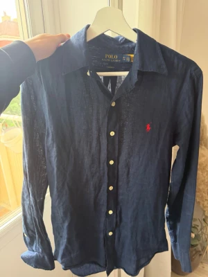 Marinblå linneskjorta från Polo Ralph Lauren - Snygg marinblå skjorta i linne från Polo Ralph Lauren med klassisk krage och vita knappar. Custom fit-modell med det ikoniska röda logobroderiet på bröstet. Perfekt för dig som gillar stilrena och tidlösa plagg.