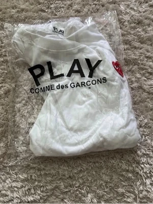 Vit t-shirt Comme des Garçons Play - Säljer en vit t-shirt från Comme des Garçons Play med svart text och det ikoniska röda hjärtat på bröstet. Klassisk passform och rund halsringning, perfekt för en stilren och trendig look.