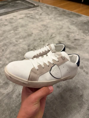 Vita Philippe Model sneakers med mockadetaljer - Snygga vita sneakers från Philippe Model med beige mockadetaljer på sidorna och vit läderöverdel.Mörkblå detalj, och knappast använda. 