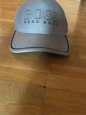 Grå keps från Hugo Boss - Snygg grå keps från Hugo Boss med broderad logga framtill. Klassisk böjd skärm och justerbart spänne bak för perfekt passform. Tillverkad i ett mjukt tyg som känns bekvämt hela dagen. Perfekt accessoar för en clean och stilren look.
